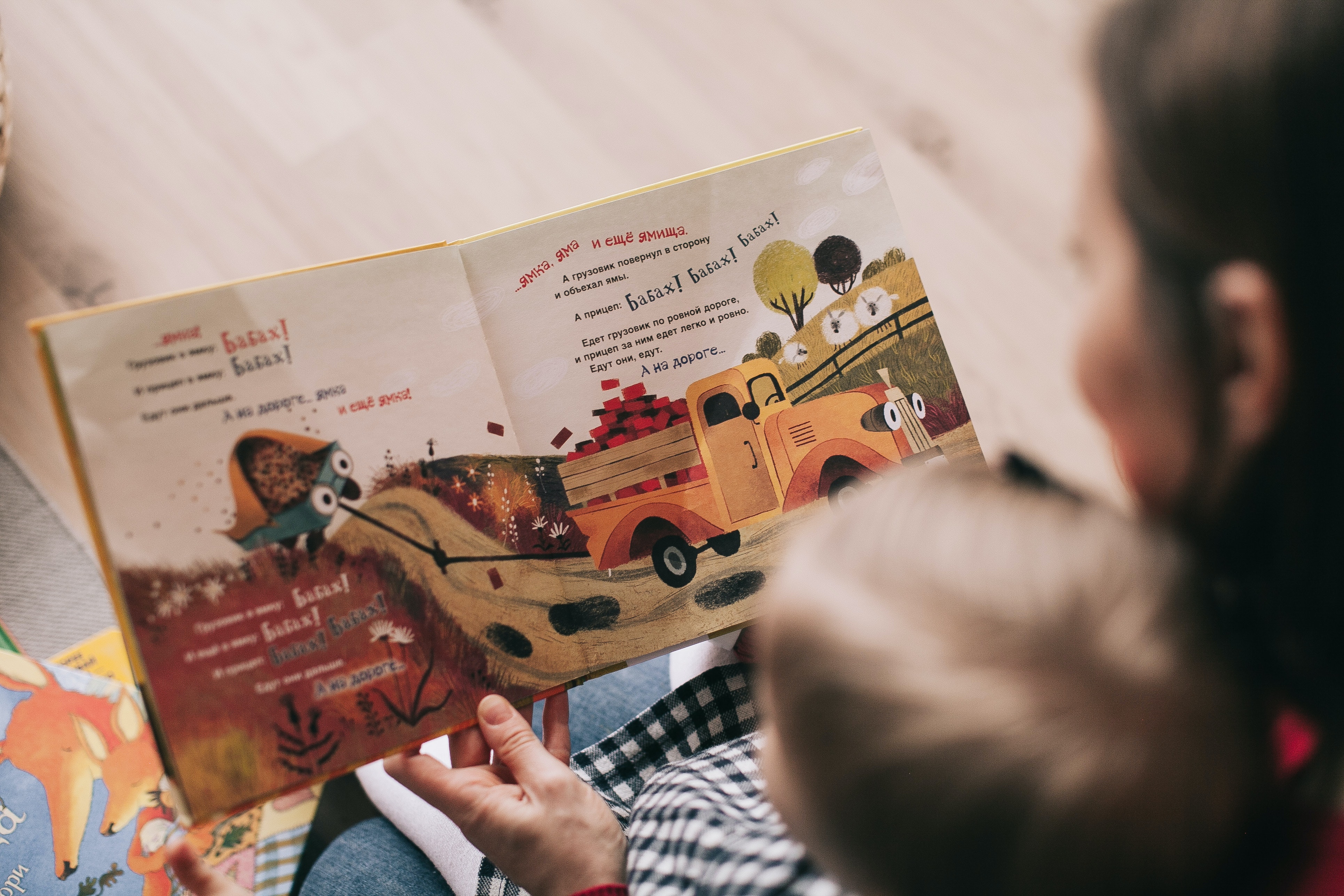 Madre sentada en un sillón, sosteniendo un libro álbum ilustrado abierto, mientras lee y muestra las coloridas imágenes a su bebé en brazos, quien la mira con atención.