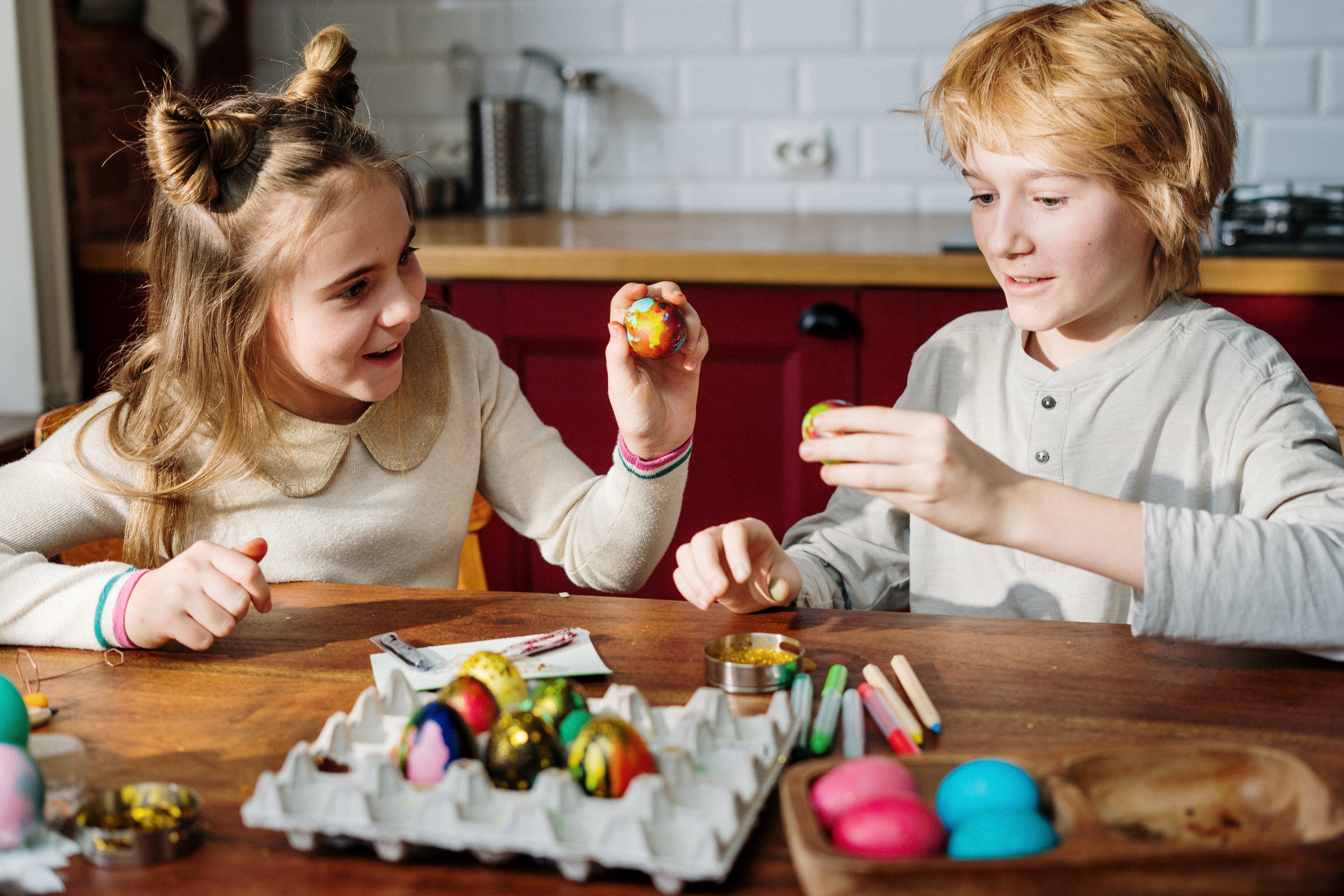Niña y niño pintando huevos de Pascua en una mesa con colores y materiales de manualidades